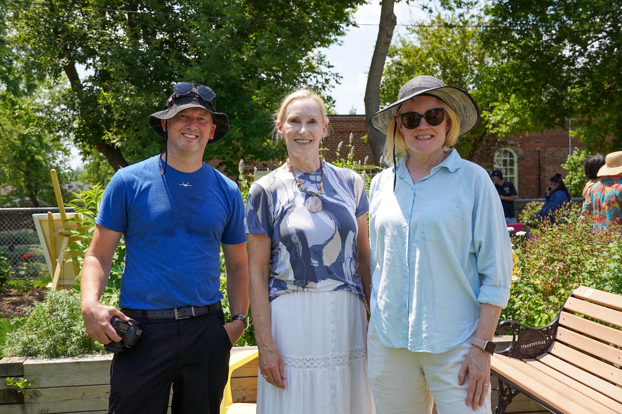 Three people wearing sun hats and casual summer clothes stand together and smile in a sunny park setting.