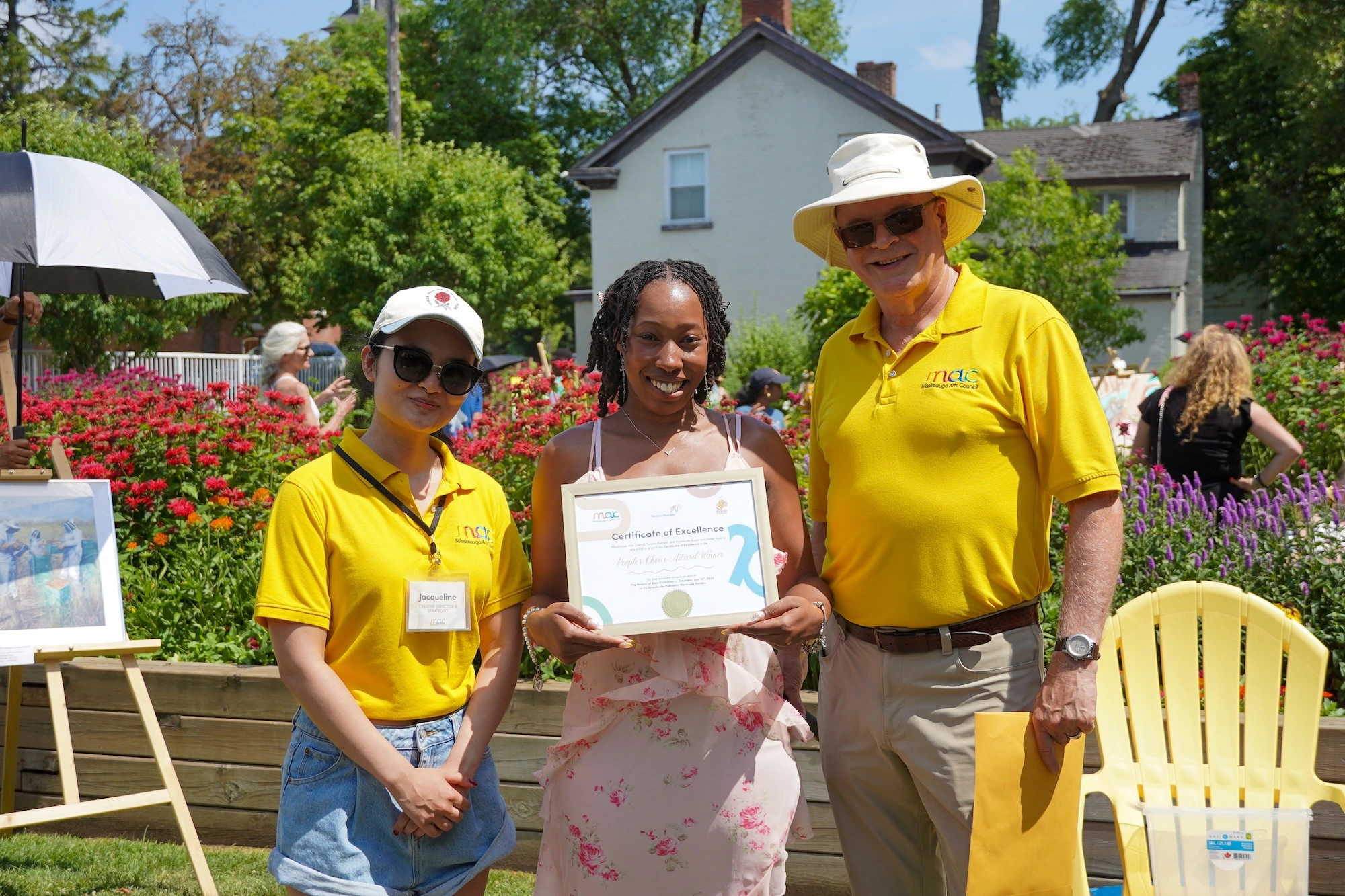 A woman holds a "Certificate of Excellence" award while standing between two event organizers in yellow shirts in a blooming garden.