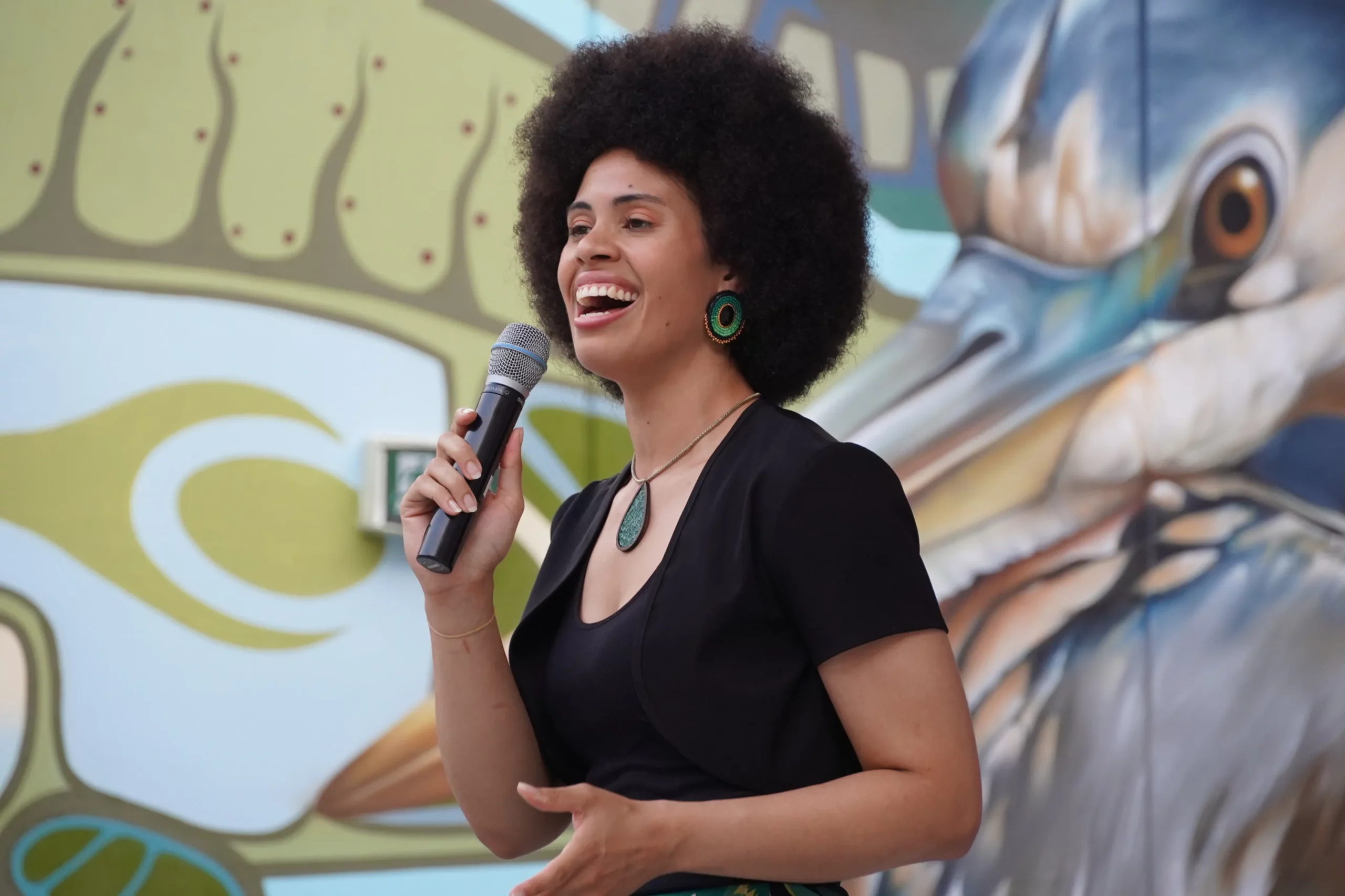A young performer sings into a microphone at an outdoor event with a vibrant mural behind them.