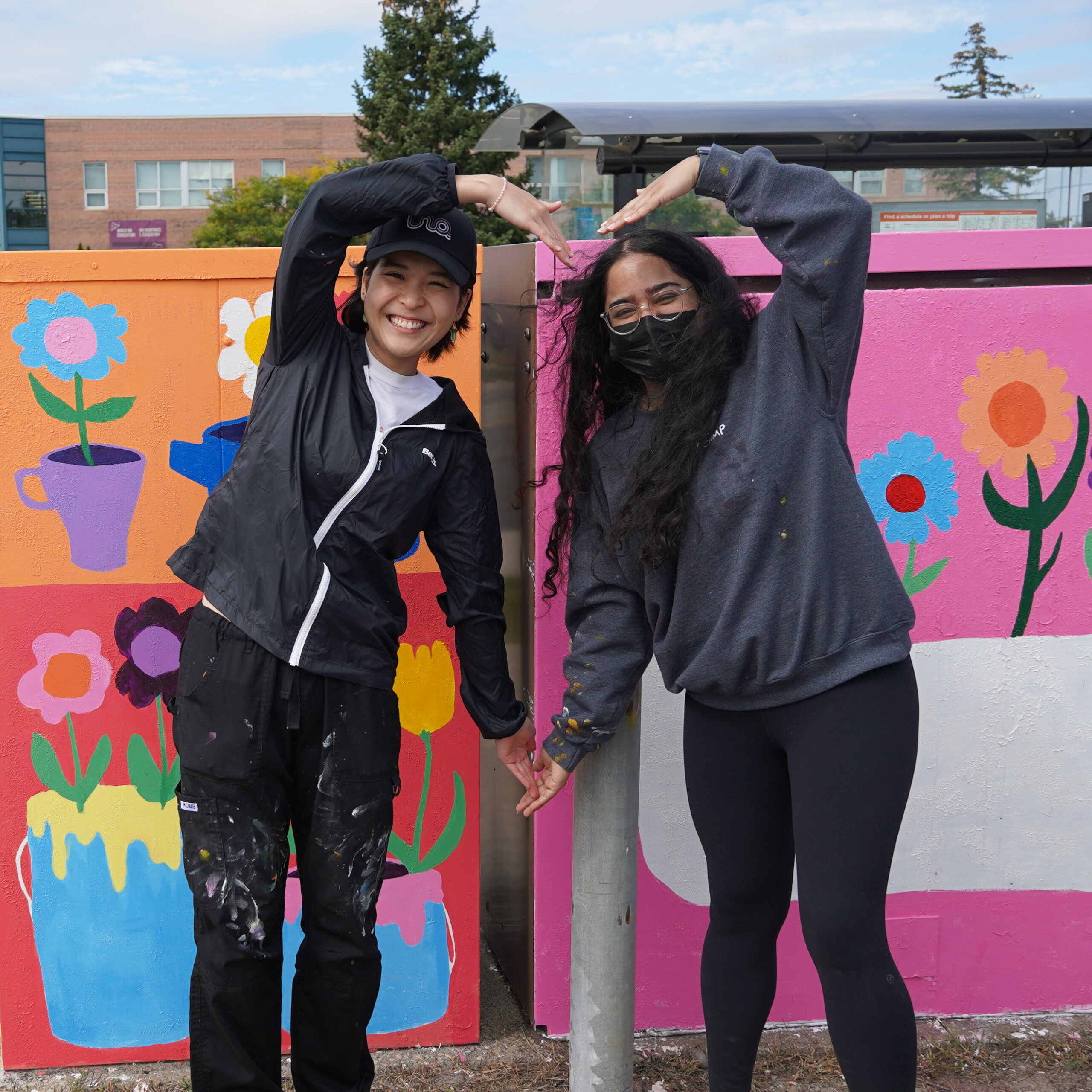 Two young women standing in front of colourful floral murals, raising their arms to form a heart shape together.
