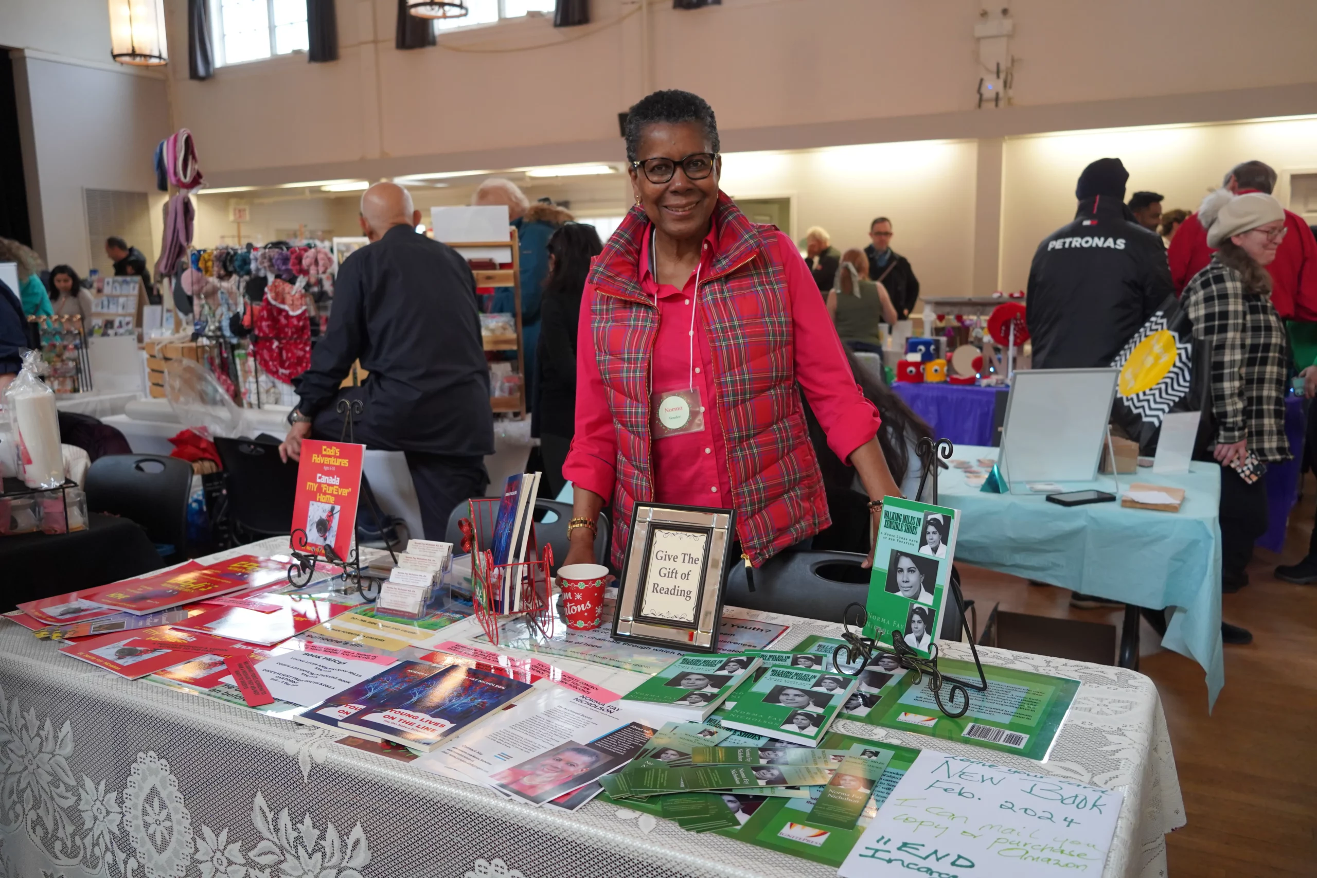 woman in a red plaid vest standing behind a table at a community market, showcasing several books.