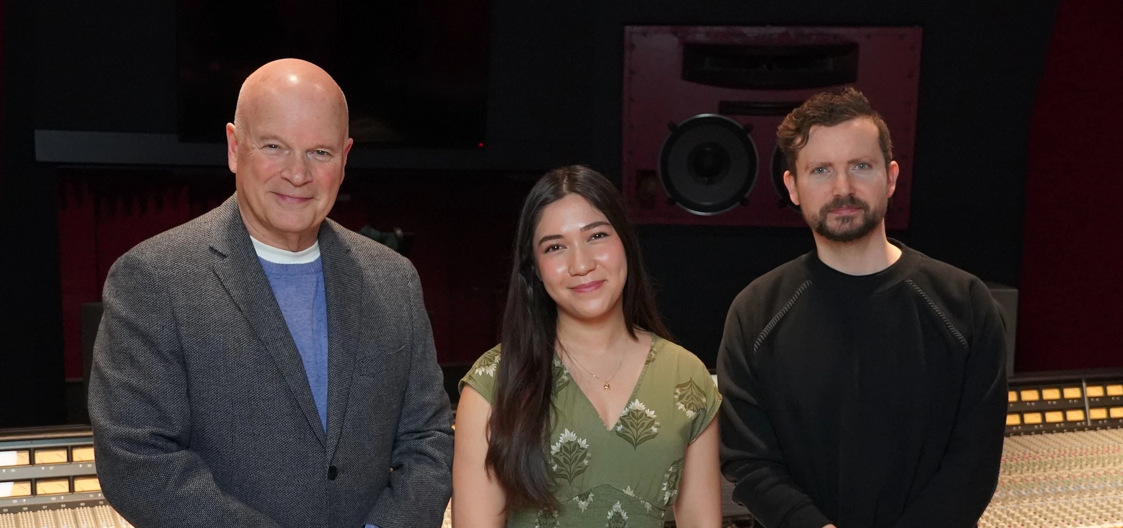 A professional portrait of an man in a grey blazer, a young woman in a green floral dress, and a man in a black sweater standing together. They are positioned in front of a wide audio mixing board within a recording studio.