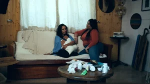 A film still showing two women sitting on a couch in a wood-paneled room. One woman holds a pillow closely while the other speaks to her; the foreground table is cluttered with pill bottles and tissues.