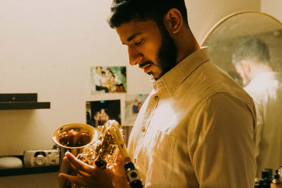 A side profile of a man with a beard, looking down thoughtfully while holding a saxophone in a dimly lit room. Behind him, personal photos and a round mirror reflecting his back are visible on the wall.