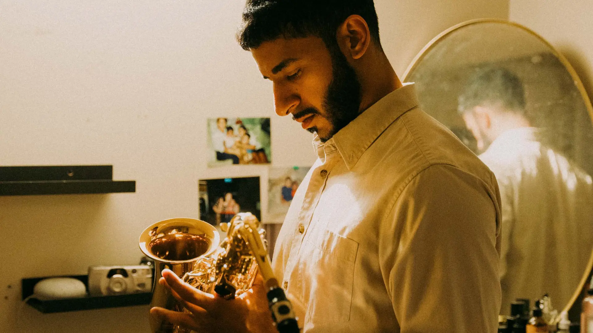 A side profile of a man with a beard, looking down thoughtfully while holding a saxophone in a dimly lit room. Behind him, personal photos and a round mirror reflecting his back are visible on the wall.