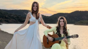 Two women posing outdoors at sunset by a lake with mountains in the background. One woman stands in a white dress while the other sits beside her playing an acoustic guitar.