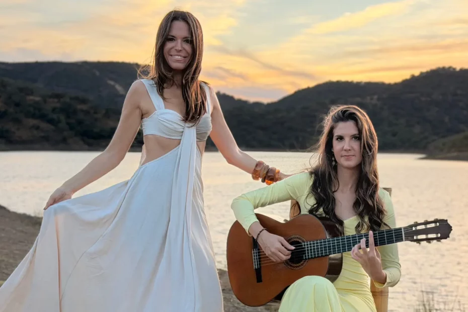 Two women posing outdoors at sunset by a lake with mountains in the background. One woman stands in a white dress while the other sits beside her playing an acoustic guitar.