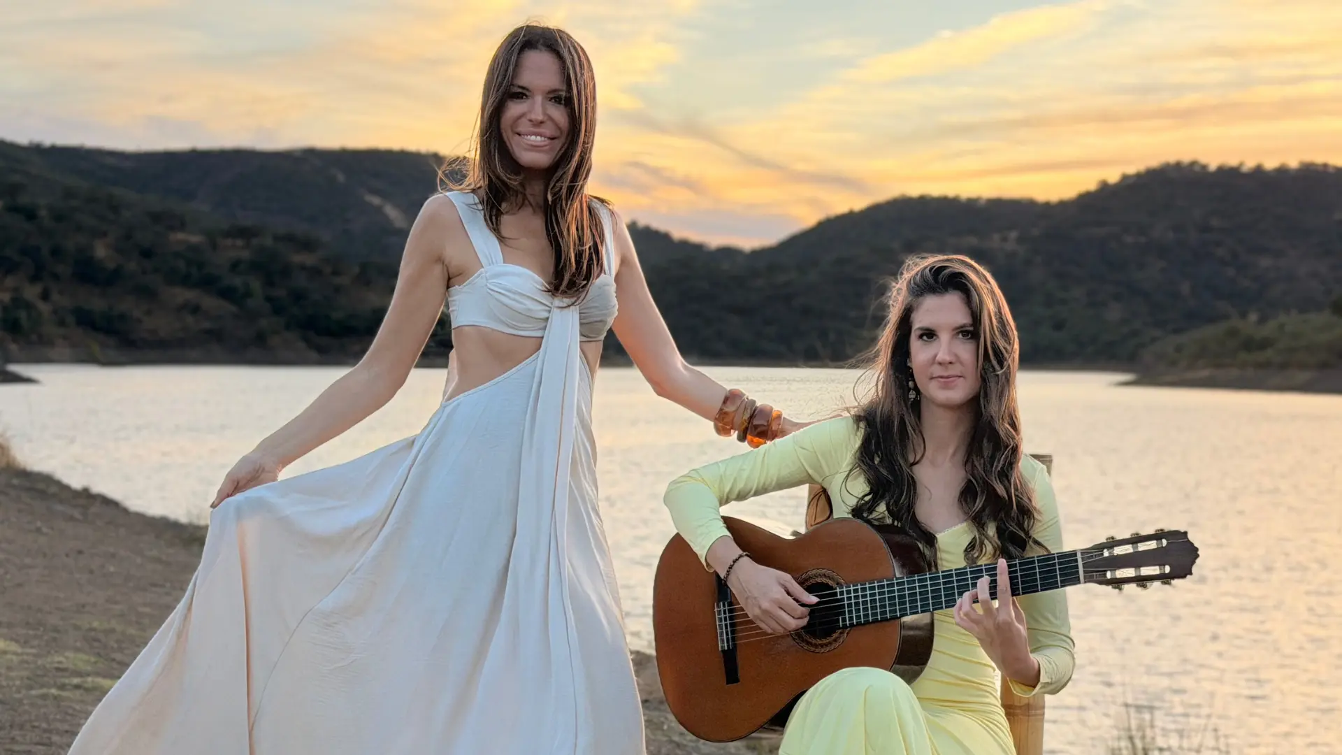 Two women posing outdoors at sunset by a lake with mountains in the background. One woman stands in a white dress while the other sits beside her playing an acoustic guitar.