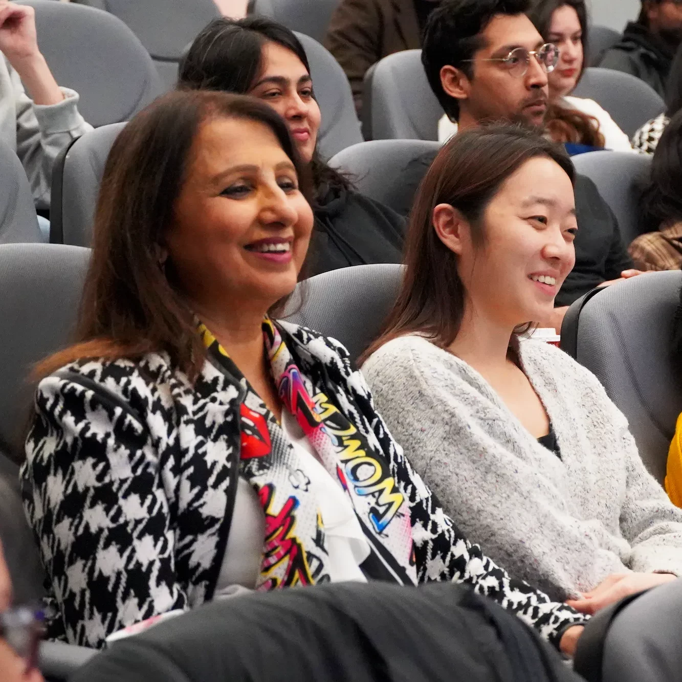 Two women sitting in a theater-style audience, smiling and looking toward a screen.