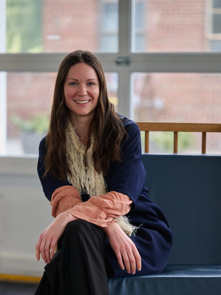 Photo of Colleen Snell seated on a blue couch in a space with natural light. Her legs are crossed, and arms resting over her knee. She has long hair, is smiling, and is wearing a blue jacket over a peach frilly shirt with a fluffy beige scarf, and black pants.