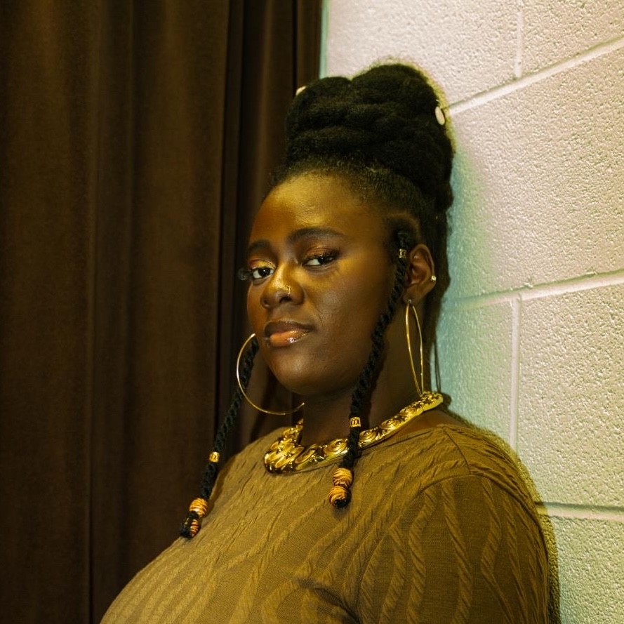 Headshot of Sewa from the shoulder up. She stands in front of a white cement wall and a brown curtain. She wears gold jewellery and a brown dress.