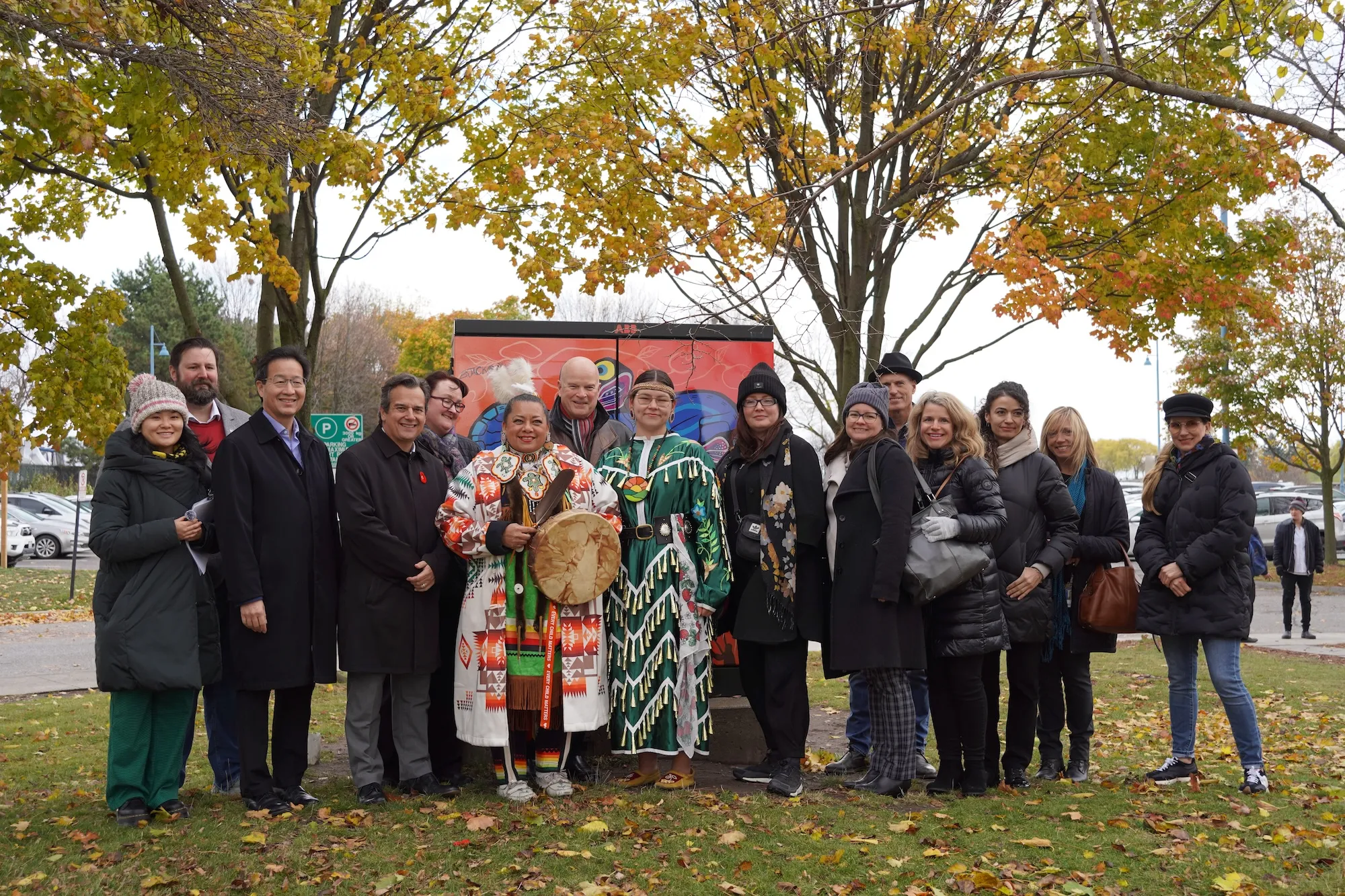 A large group of people posing outdoors in front of a colourful vinyl-wrapped utility box mural. Two women in the centre are wearing traditional Indigenous regalia, with one holding a ceremonial drum.