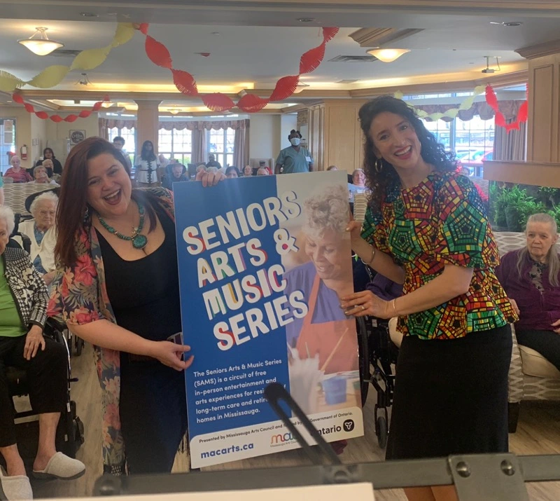 Two women smile and hold up a large blue "Seniors Arts & Music Series" poster while posing in front of an audience.