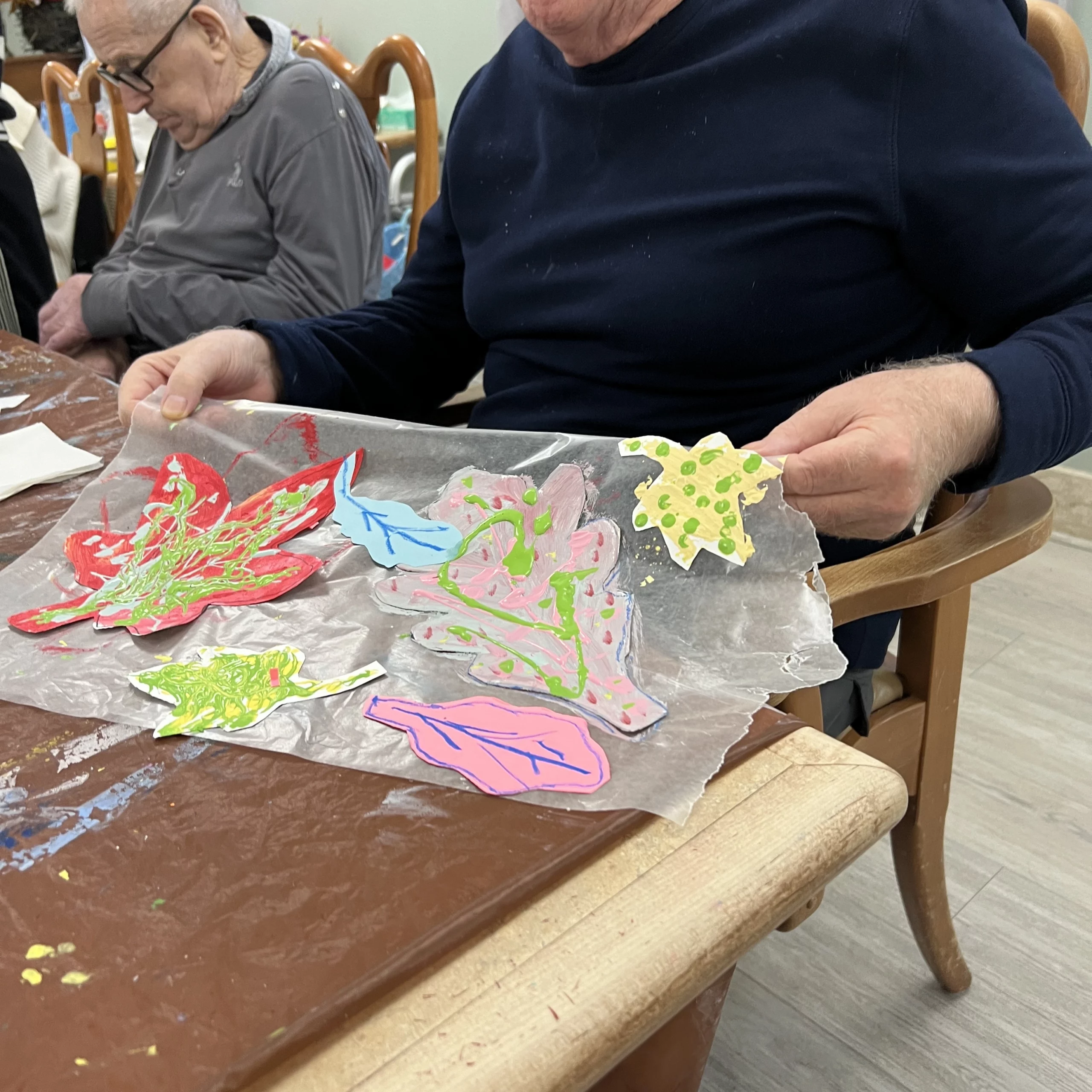 Close-up of a senior resident’s hands holding a colorful autumn leaf art project made with paint and paper.