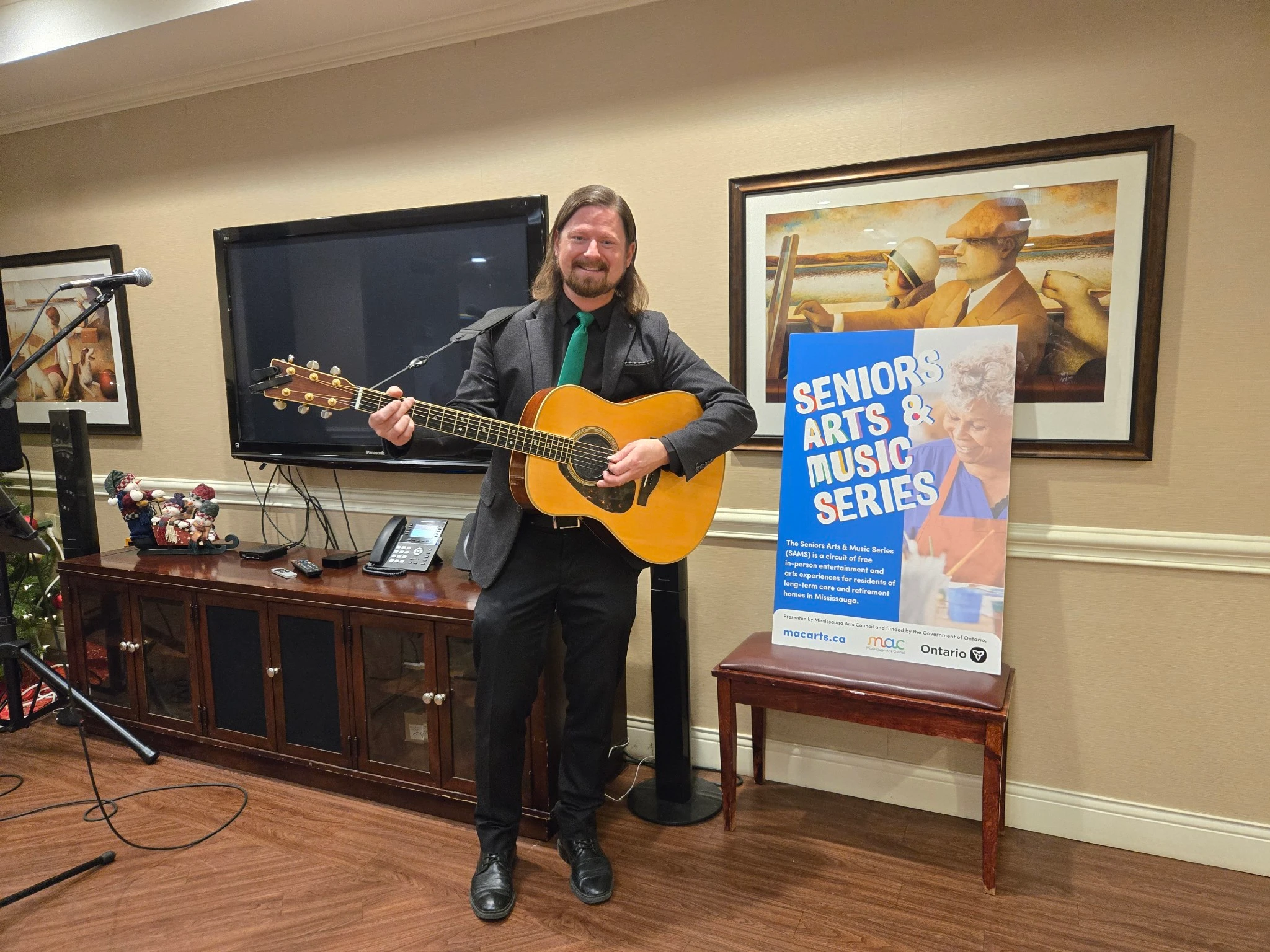 A smiling musician with long hair and a green tie plays an acoustic guitar next to a large event poster in a cozy lounge.