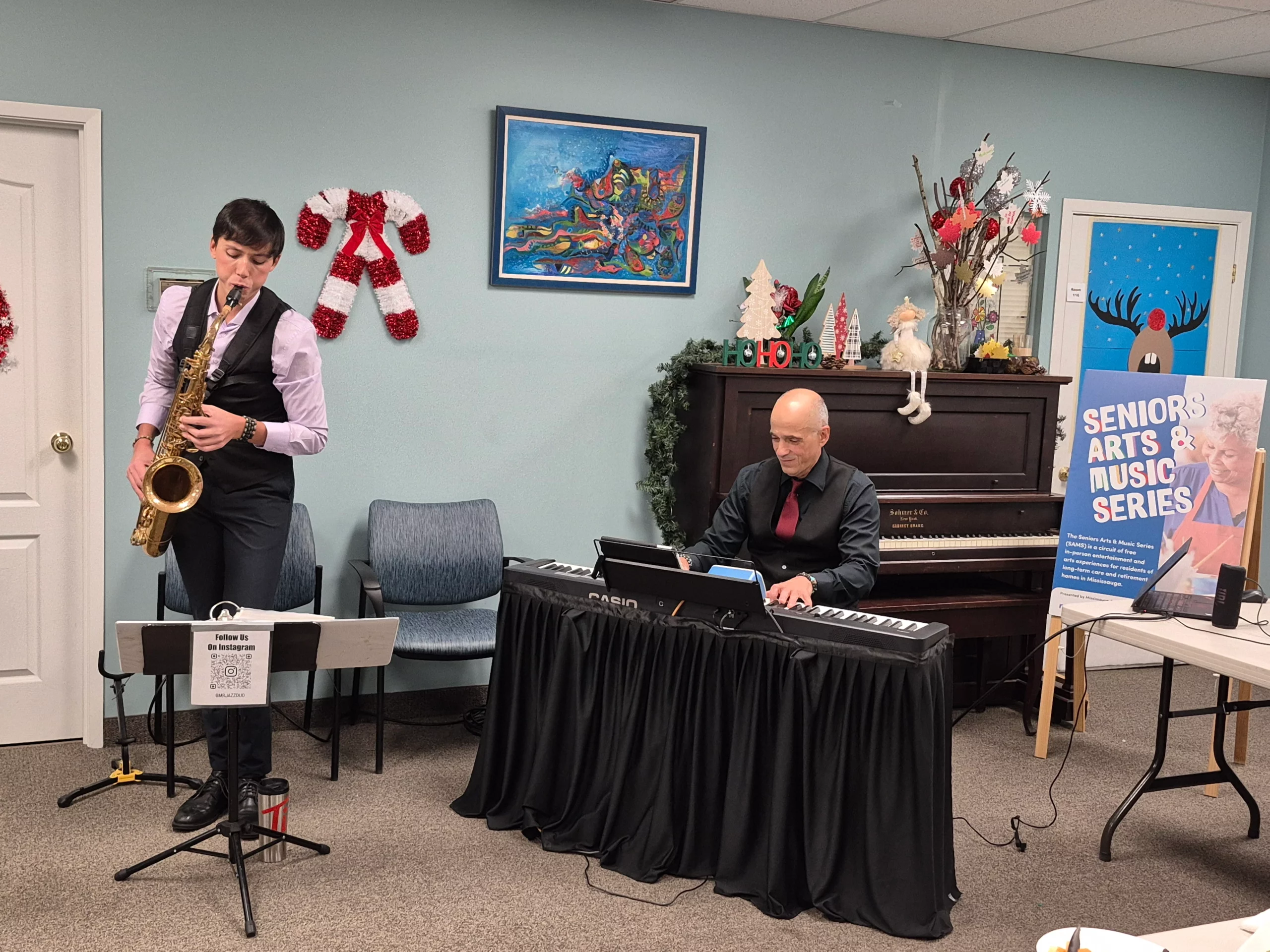 A saxophonist and a pianist in matching black vests perform in a room decorated for the holidays with a "Seniors Arts & Music Series" poster nearby.