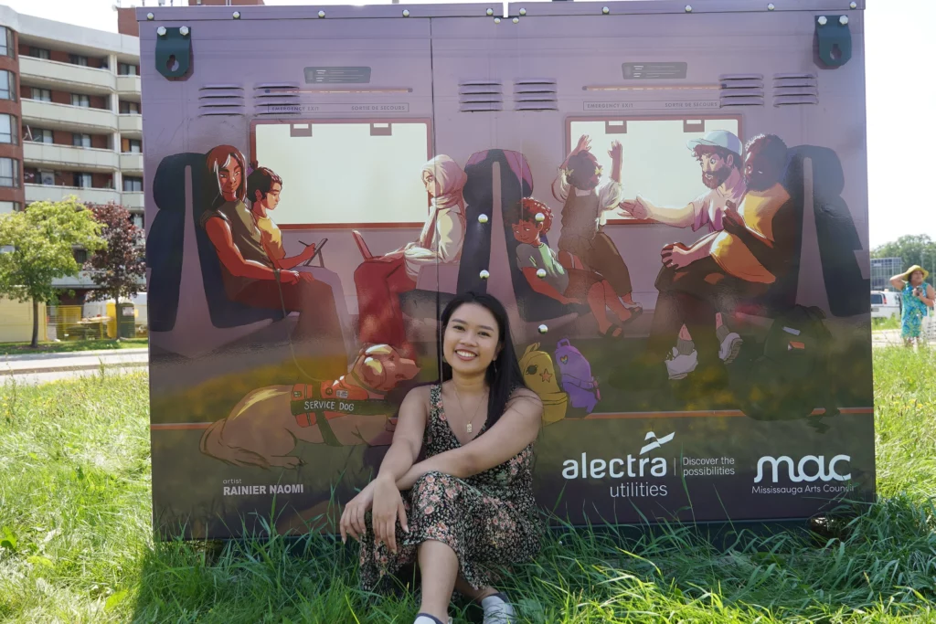 Artist Rainier Naomi sits in the grass in front of her mural on a utility box, which depicts diverse people and a service dog riding in a GO train.