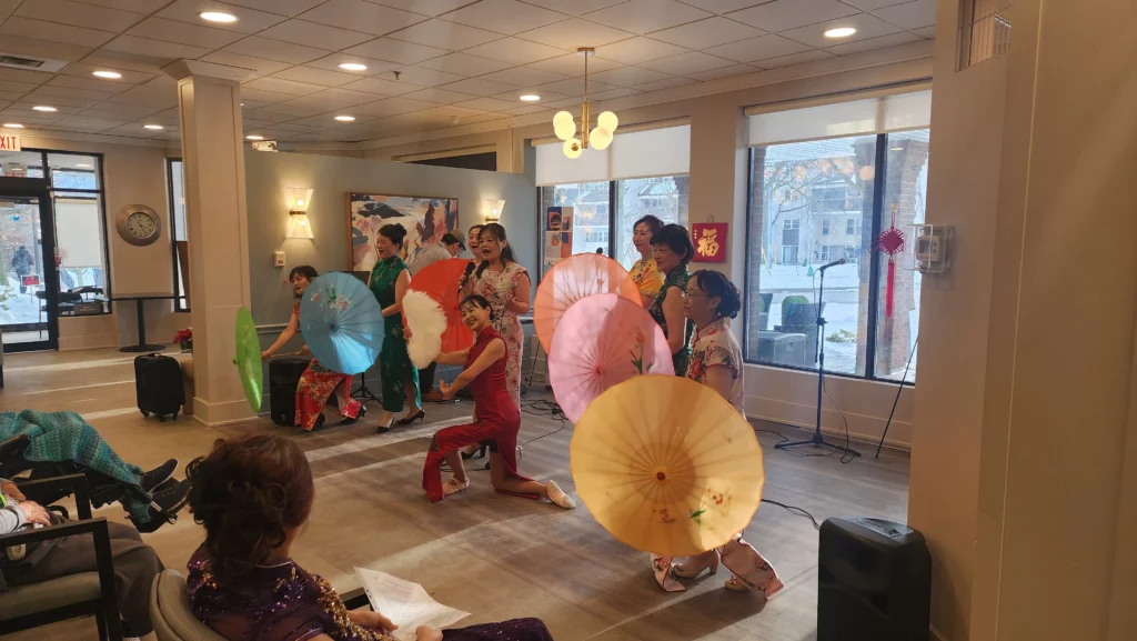 A group of women in traditional floral dresses perform a dance with colorful paper umbrellas in a sunlit community room.