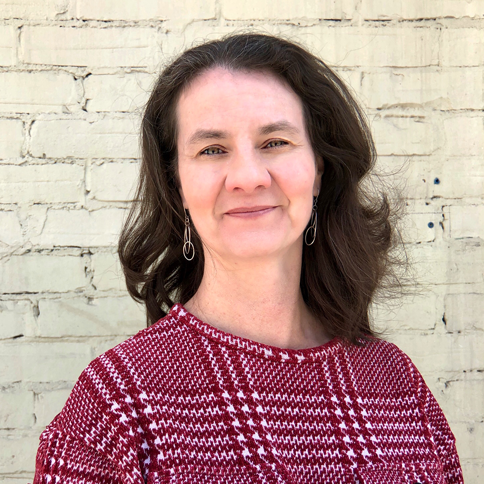 Headshot of Maura Broadhurst. She is standing against a white brick backdrop, with her brown hair down, and a plaid burgundy and white sweater.