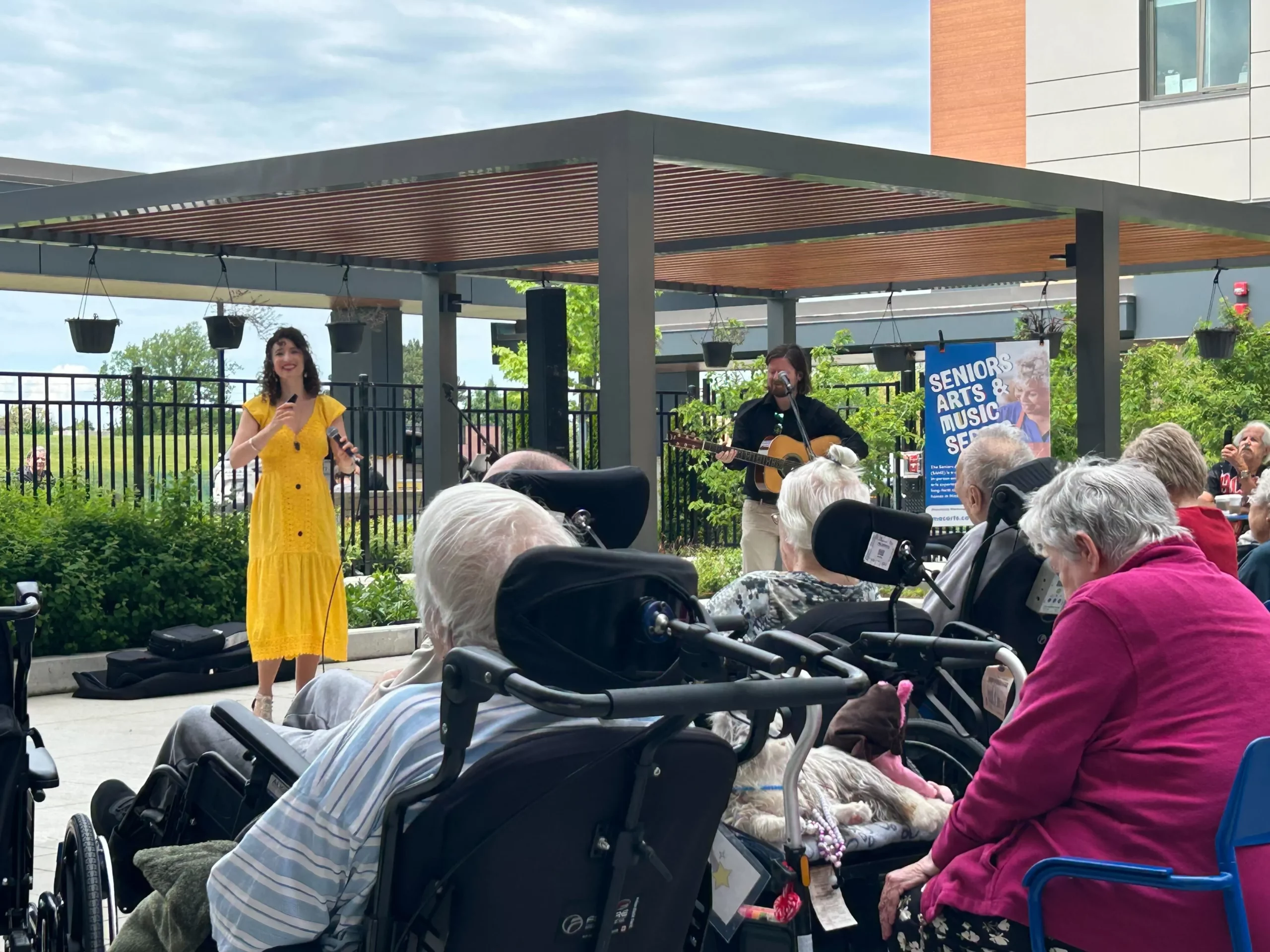 A woman in a bright yellow dress and a man with an acoustic guitar perform under an outdoor pergola for a seated audience of seniors.
