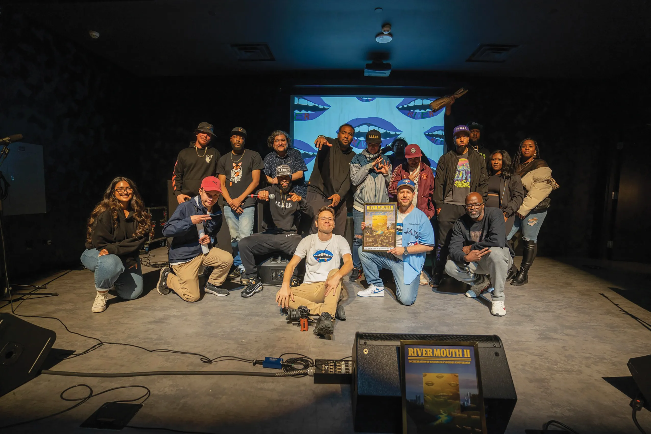 A large group of diverse artists and organizers posing together on a stage. Two men in the centre hold a framed "River Mouth II" event poster, and the background screen features a blue lip graphic.