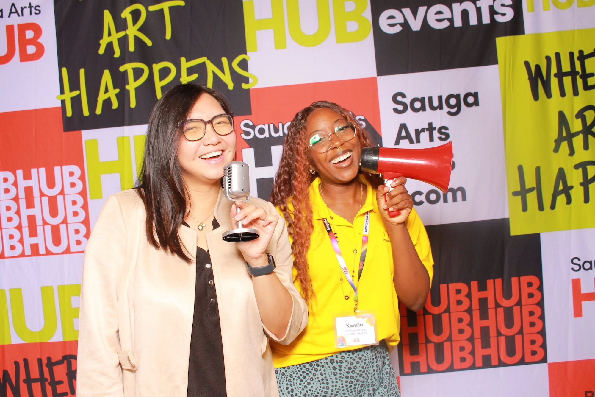 Two women laughing in a photobooth with a "Sauga Arts HUB" backdrop; one holds a vintage microphone and the other holds a red megaphone.