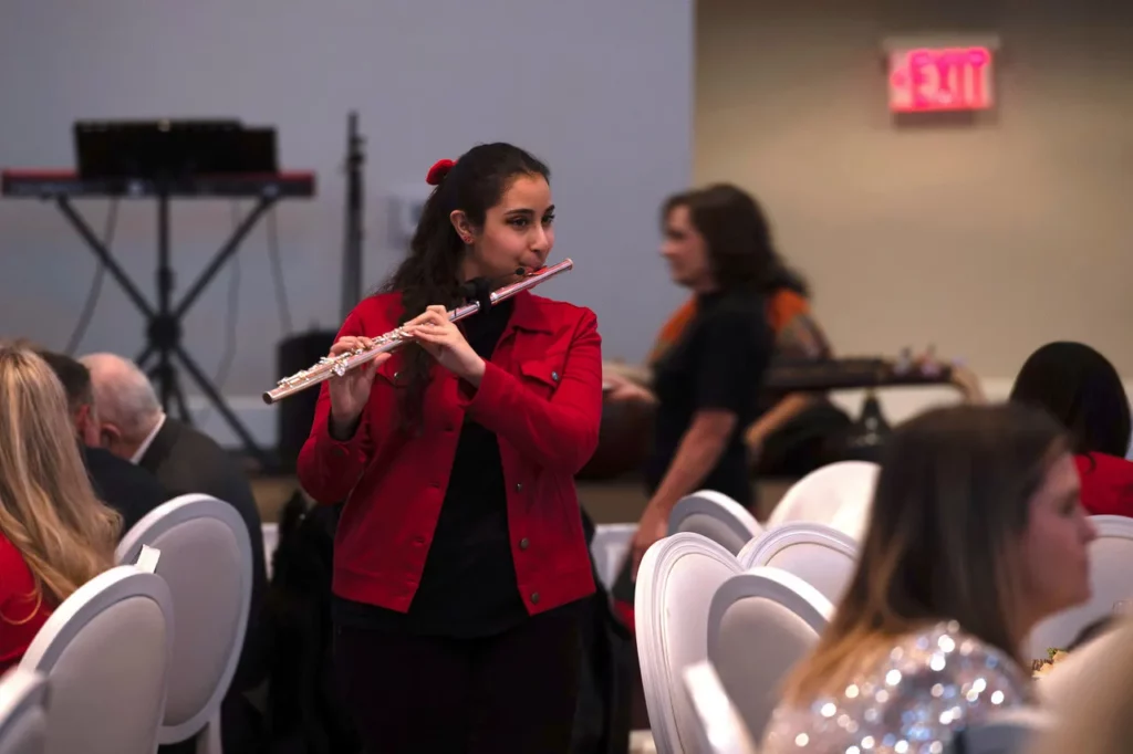 A young woman in a red jacket plays a silver flute while standing in a banquet hall among seated guests.
