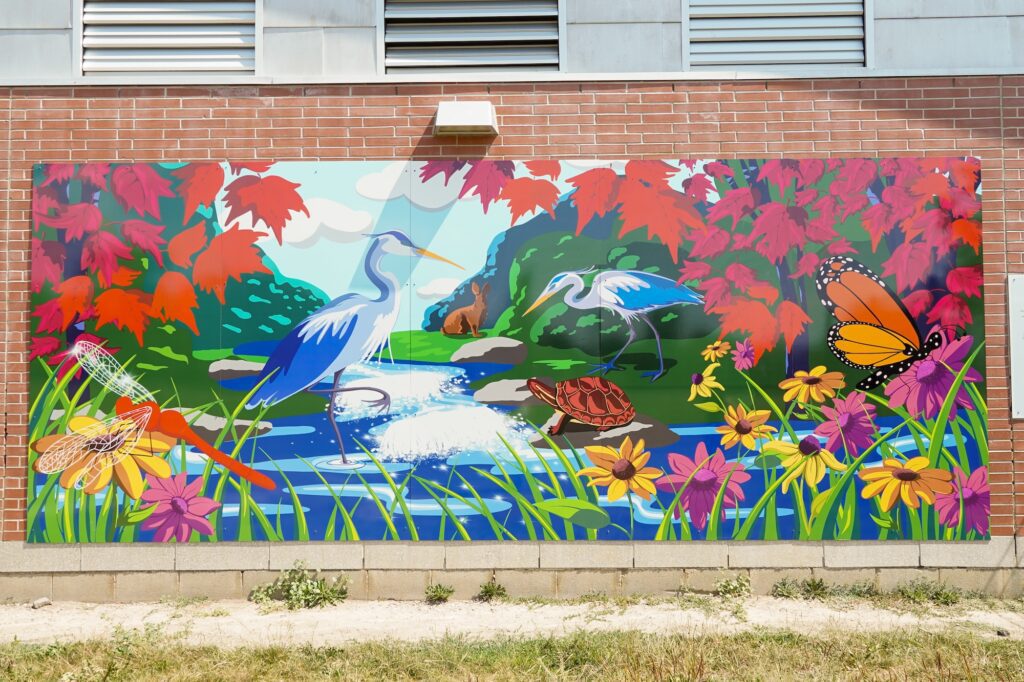 A vinyl mural panel on the side of a brick wall. The colourful mural depicts a scene with a realistic and brightly coloured park with a river running in the middle. With dense foliage, animals like a crane, rabbit, and turtle are visible.