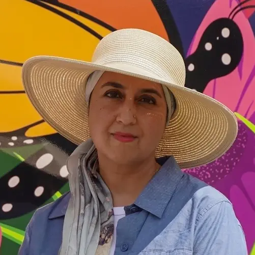 Two women standing side-by-side in front of a vibrant, colourful mural featuring a large butterfly and flowers. The woman on the left wears sunglasses and a floral tank top, while the woman on the right wears a wide-brimmed straw hat and a blue button-down dress.