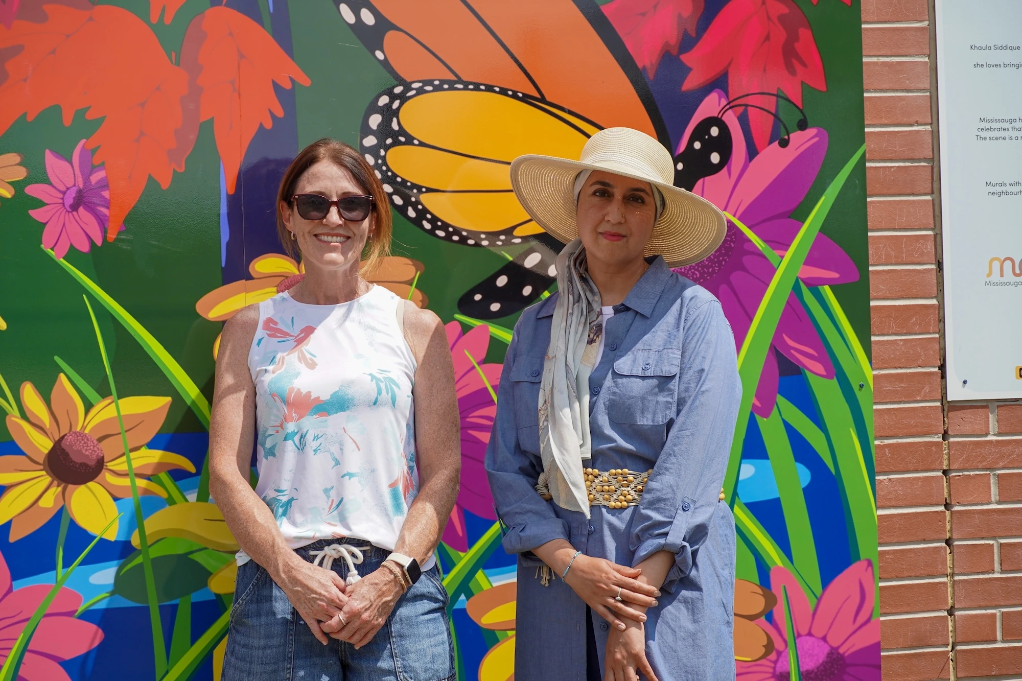 Two women standing side-by-side in front of a vibrant, colourful mural featuring a large butterfly and flowers. The woman on the left wears sunglasses and a floral tank top, while the woman on the right wears a wide-brimmed straw hat and a blue button-down dress.