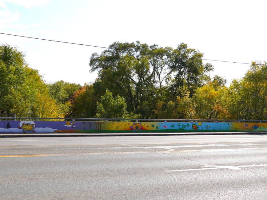 A large-scale hand-painted mural on a cement bridge guardrail. On it is an Indigenous style mural that depicts different seasons on four panels, each with a turtle that embodies the season, as well as other local plant and animal species.