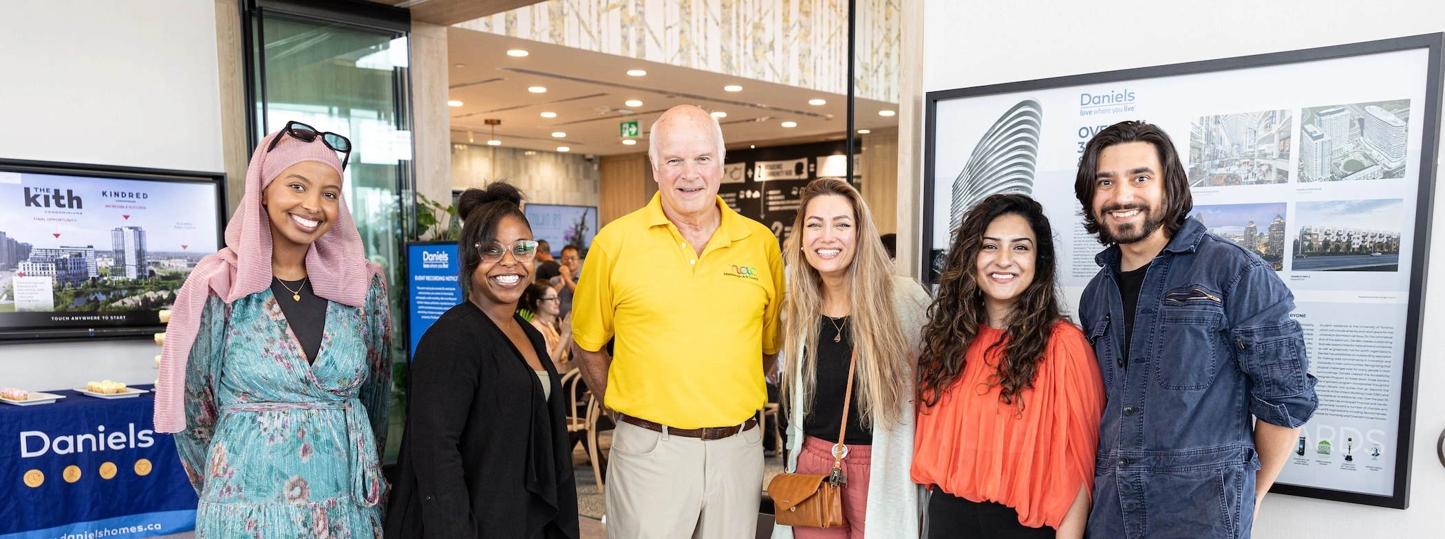 A smiling group of six people standing together in a brightly lit modern lobby. They are posed in front of a wall display featuring architectural renderings and the "Daniels" brand name.