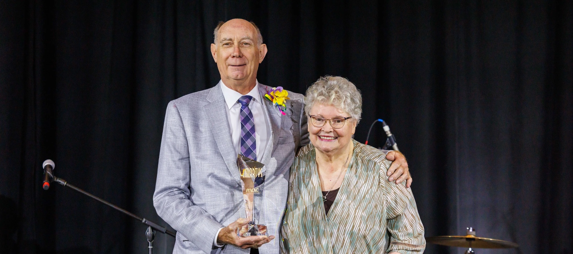 A man in a light grey suit and a woman in a patterned blouse smiling together on stage. The man is holding a glass MARTY award trophy.