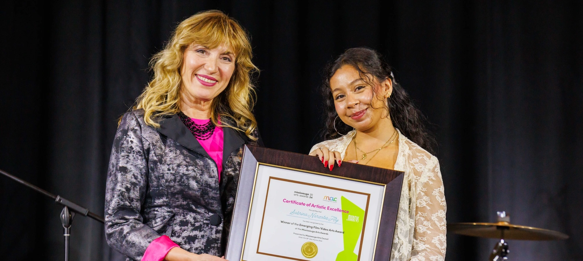 Two women smiling on stage while holding a framed "Certificate of Artistic Excellence" from the Mississauga Arts Council.