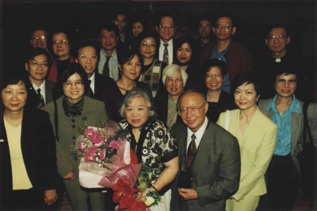 A large group of people are photographed from a high angle in a dark space. In front, a woman (Bernice Kwong) holds a bouquet. 