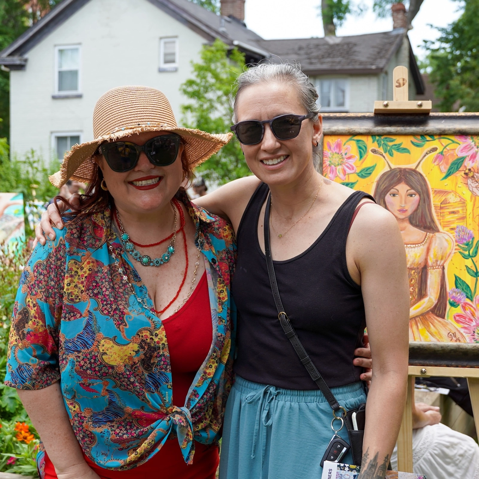 Two women smiling for a photo outdoors at an art event. The woman on the left is wearing a straw hat and a colourful patterned shirt over a red top. The woman on the right is wearing sunglasses and a black tank top. Beside them is a colourful painting on an easel featuring a woman and floral/bee designs.