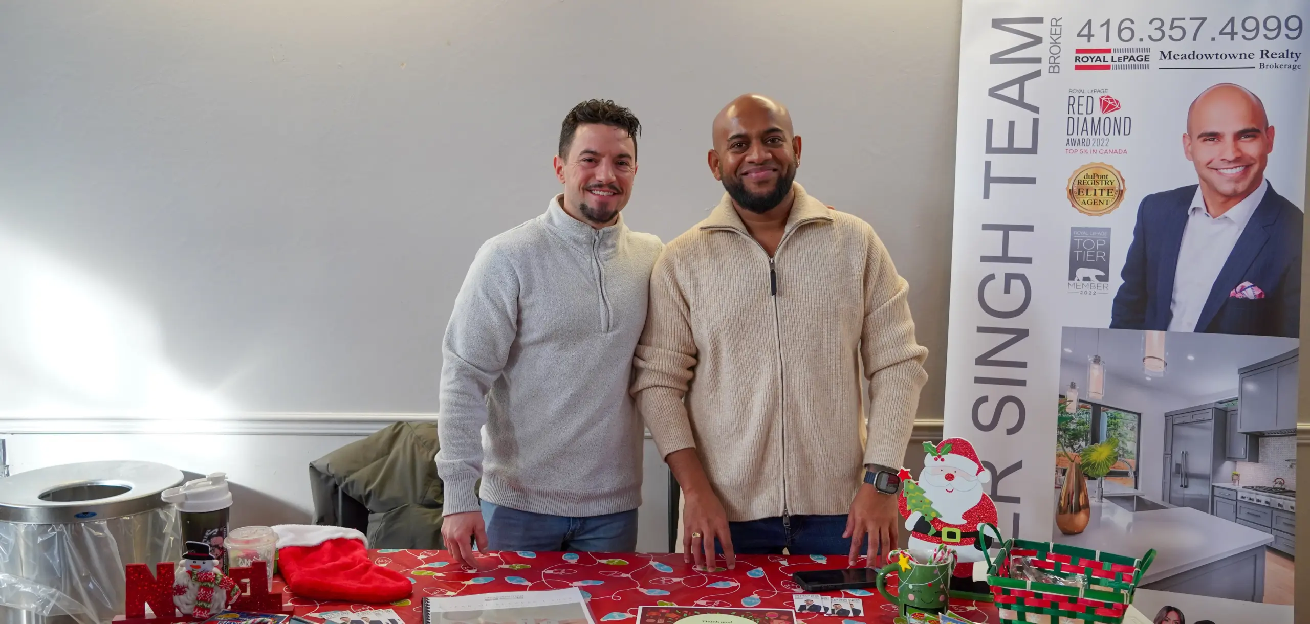Two men smiling while standing behind a table decorated for the holidays with a red tablecloth, a Christmas stocking, and a small Santa figure. To their right is a large promotional banner for "The Pinder Singh Team" real estate.