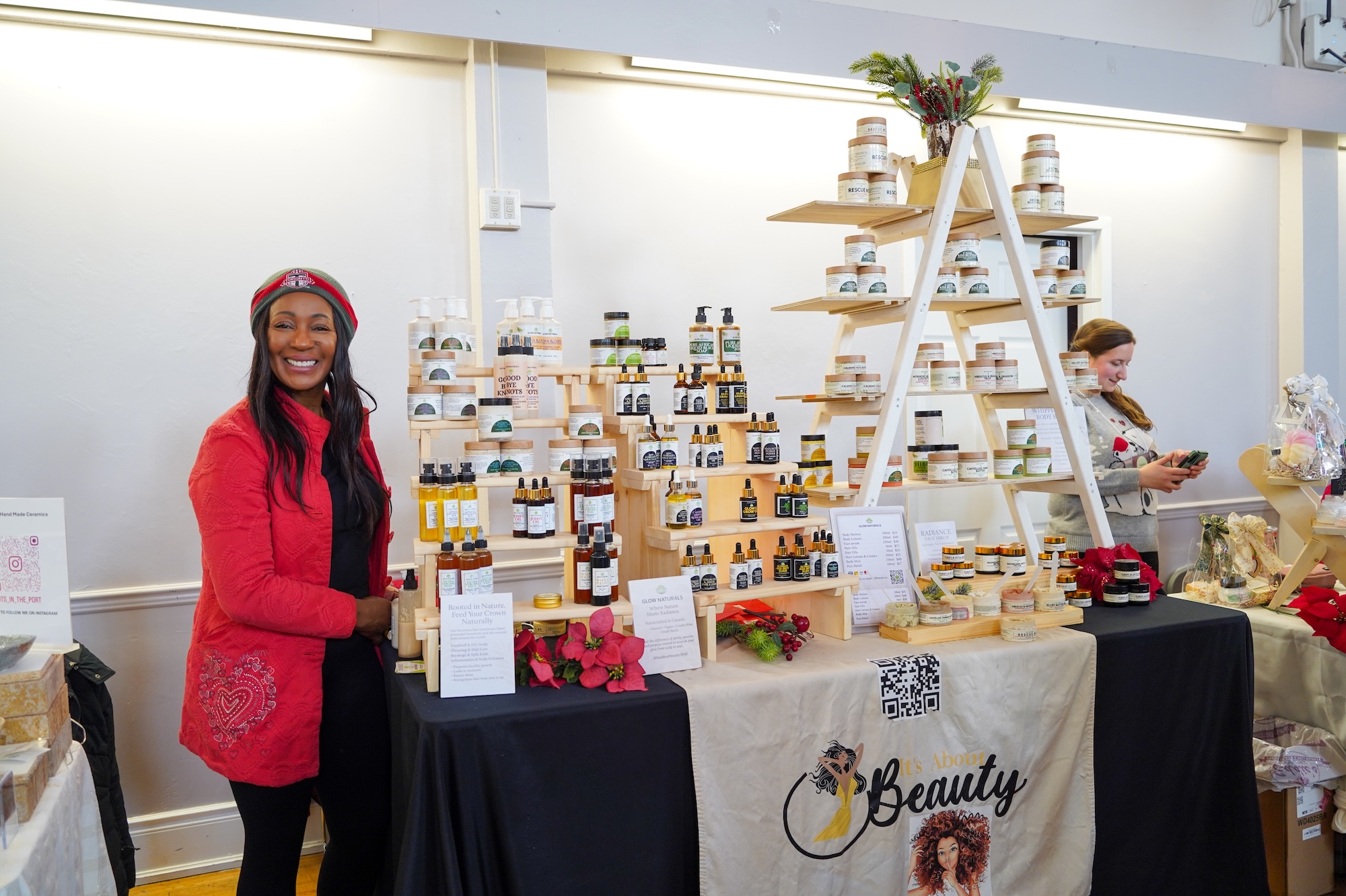 A woman in a red coat and a grey and red beanie stands proudly next to her "It’s About Beauty" vendor booth. The display features a large white A-frame shelf and smaller wooden risers packed with various skincare bottles, jars, and oils. A woman in a red coat and a grey and red beanie stands proudly next to her "It’s About Beauty" vendor booth. The display features a large white A-frame shelf and smaller wooden risers packed with various skincare bottles, jars, and oils.