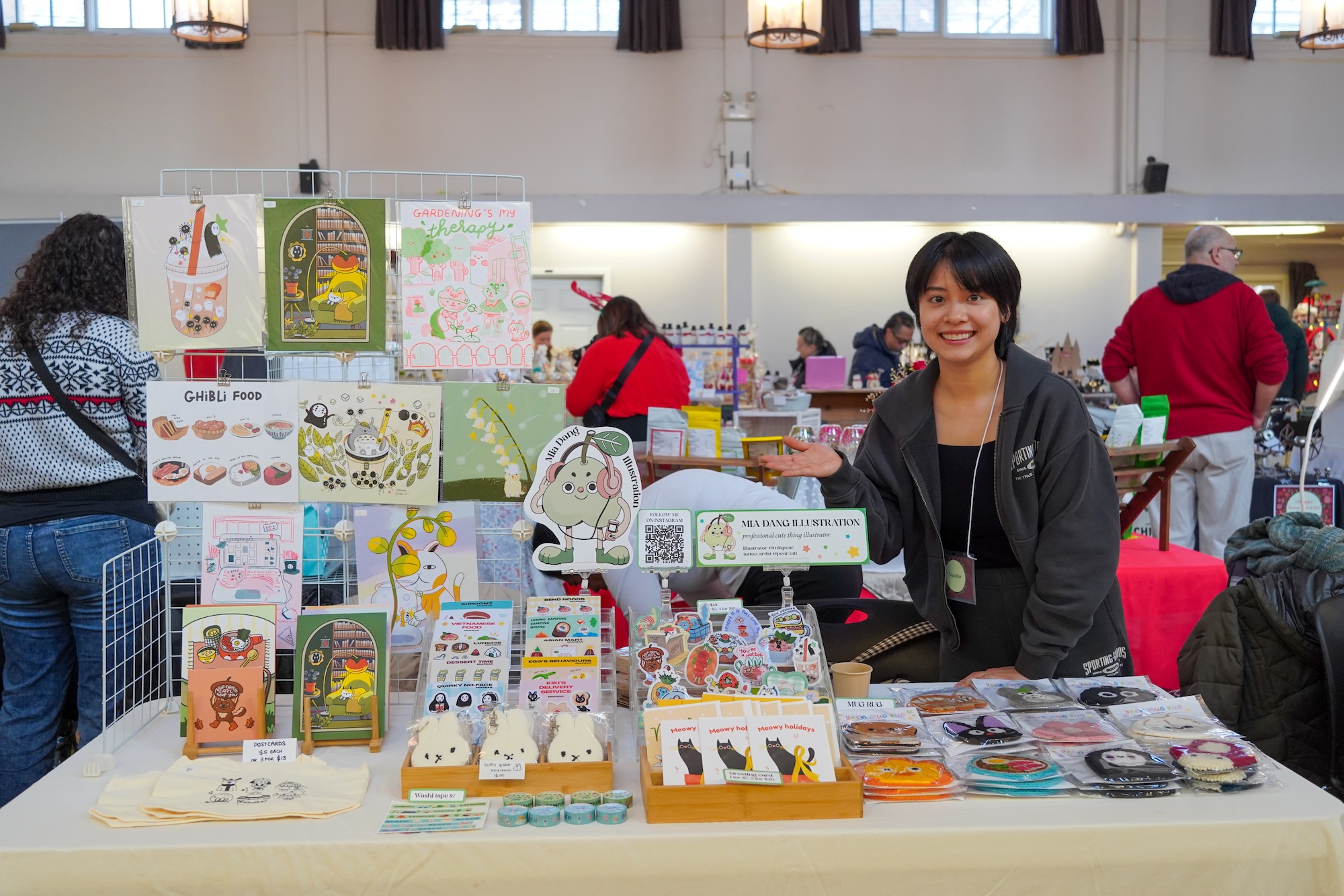 A young person gestures toward their "Mia Dang Illustration" booth at a craft fair. Their table is covered with colourful stickers, postcards, washi tape, and art prints featuring whimsical characters and food illustrations. A young person gestures toward their "Mia Dang Illustration" booth at a craft fair. Their table is covered with colourful stickers, postcards, washi tape, and art prints featuring whimsical characters and food illustrations.