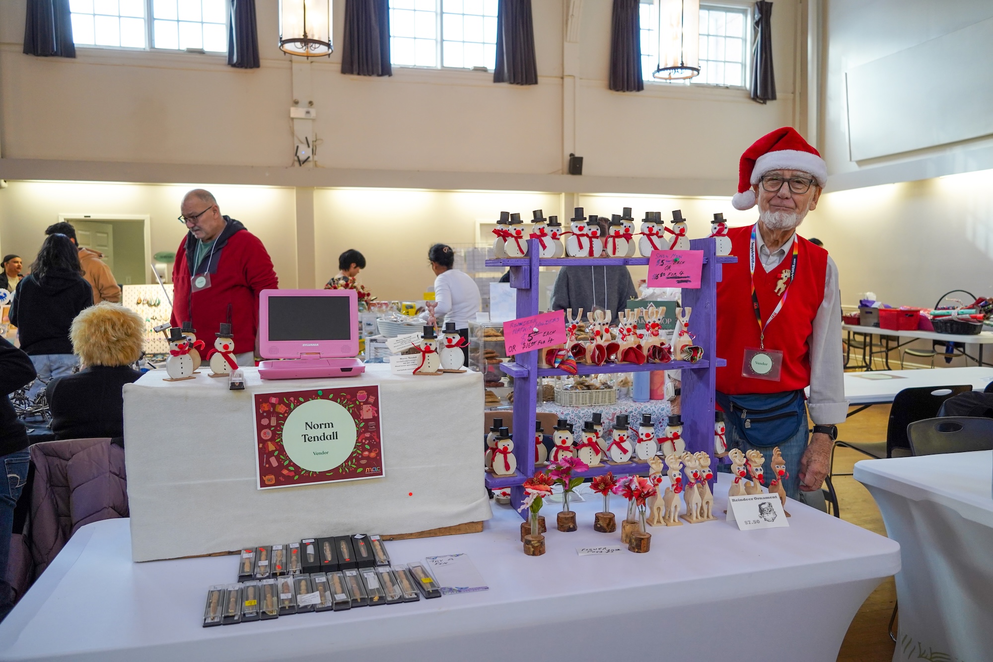 A man in a red vest and Santa hat stands behind a vendor table labeled "Norm Tendall." The booth displays rows of small, handmade wooden snowman and reindeer figurines on a purple wooden shelf, alongside boxed pens or tools on the table's surface. A man in a red vest and Santa hat stands behind a vendor table labeled "Norm Tendall." The booth displays rows of small, handmade wooden snowman and reindeer figurines on a purple wooden shelf, alongside boxed pens or tools on the table's surface.