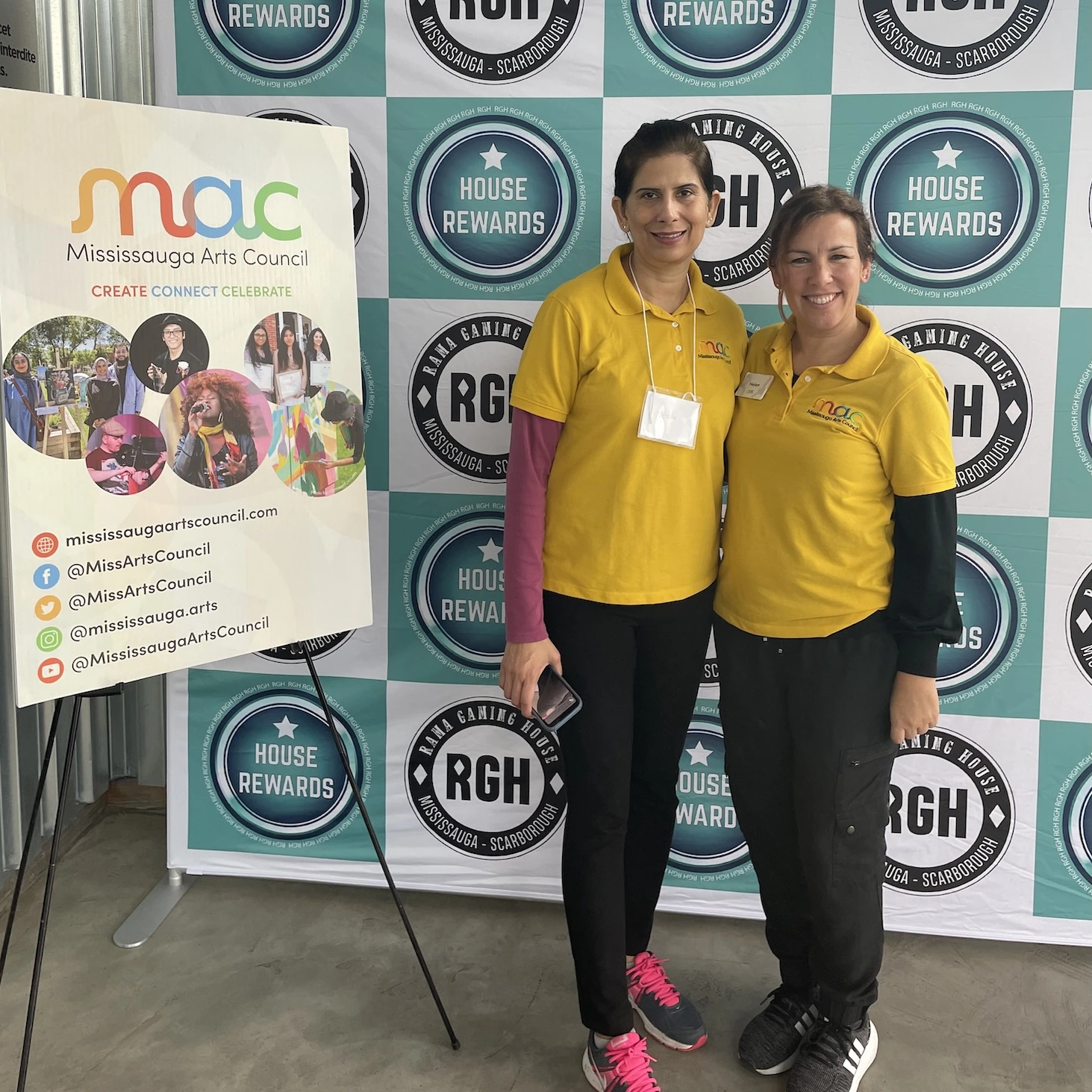 Two volunteers, Helen and Neelu, wearing yellow Mississauga Arts Council polo shirts, stand smiling in front of a Rama Gaming House branded backdrop and a MAC poster.