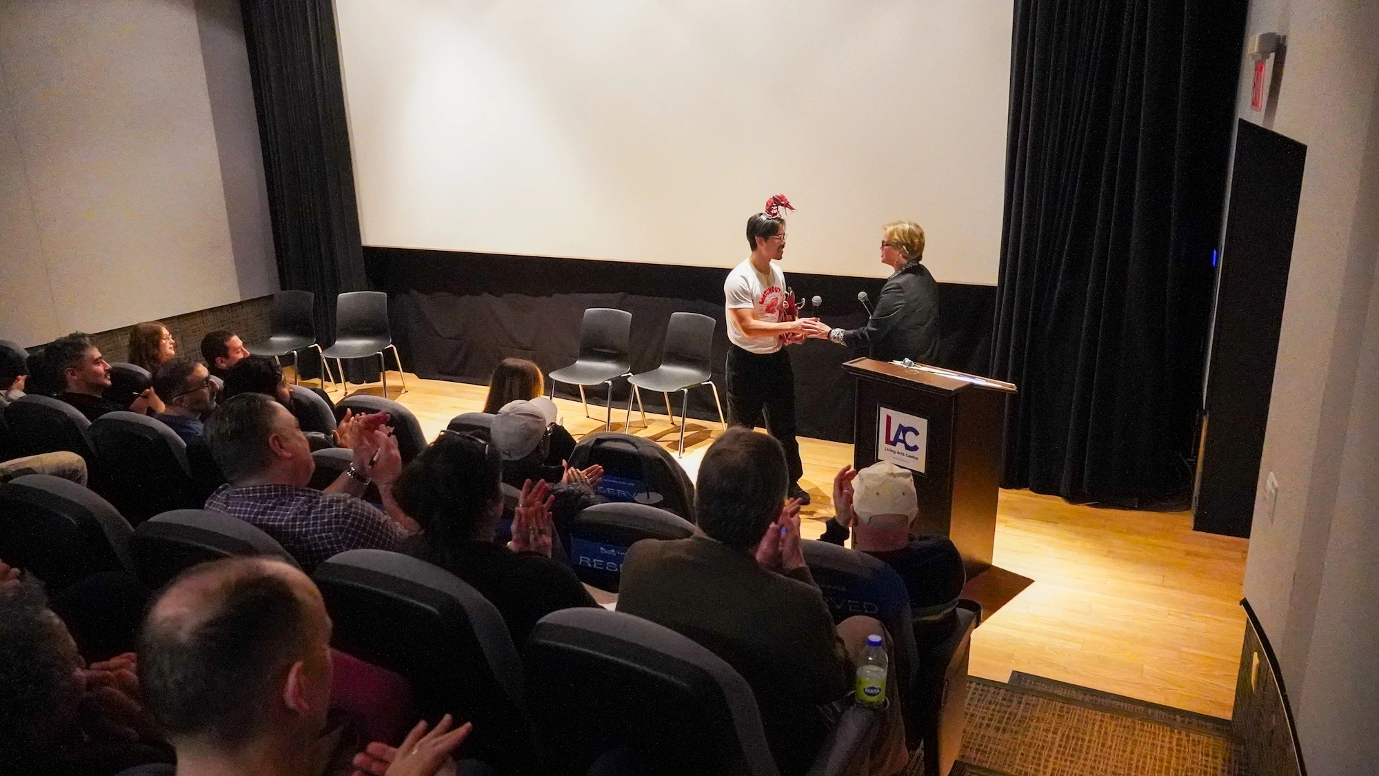 From the perspective of the audience, a man on a small stage in front of a white screen greets a woman. The room is a theatre with black curtains and wooden flooring. From the perspective of the audience, a man on a small stage in front of a white screen greets a woman. The room is a theatre with black curtains and wooden flooring.