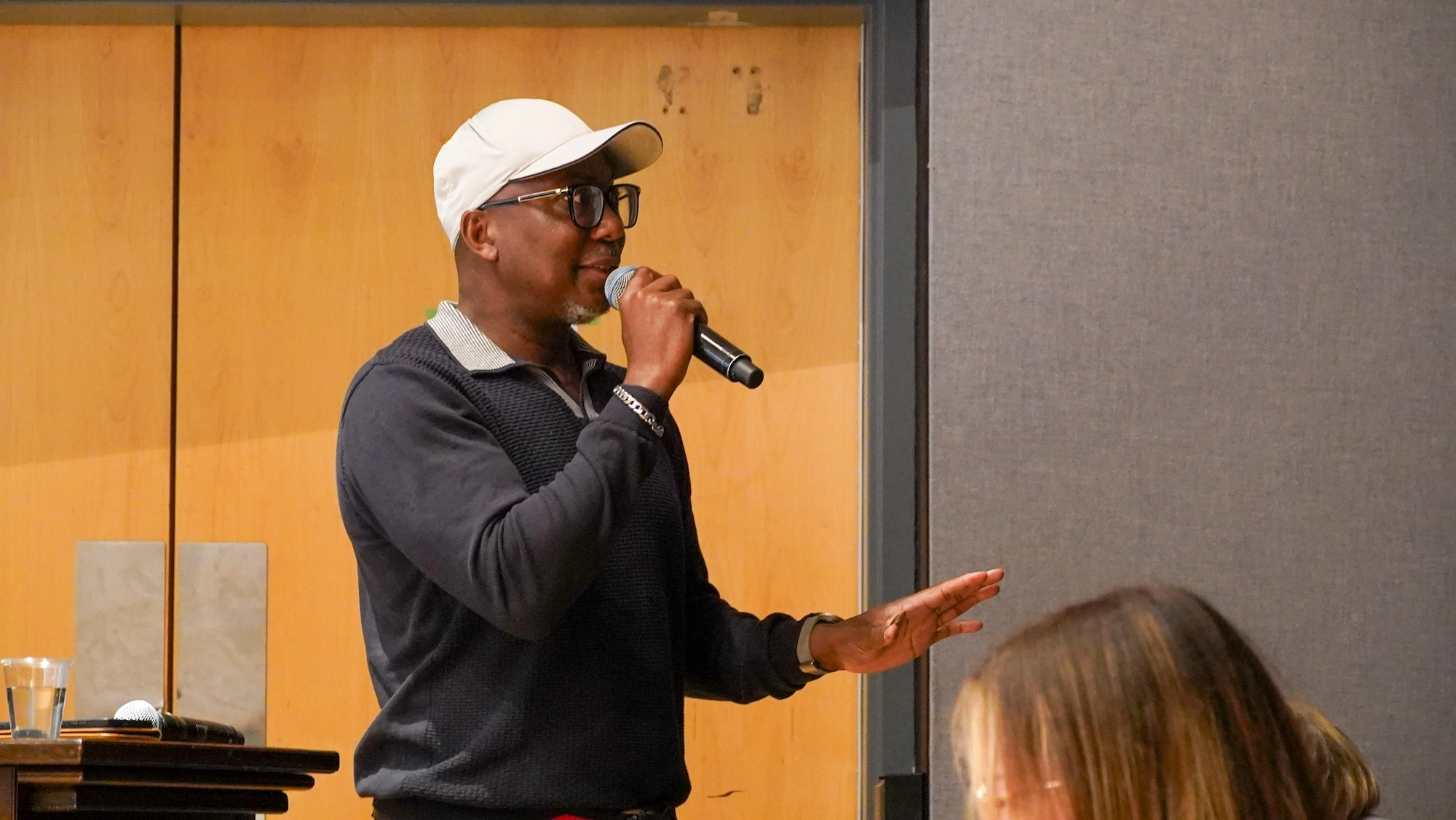 A medium shot of a man wearing a white baseball cap, glasses, and a navy polo shirt holding a microphone and speaking. He has one hand raised as if making a point. A medium shot of a man wearing a white baseball cap, glasses, and a navy polo shirt holding a microphone and speaking. He has one hand raised as if making a point.