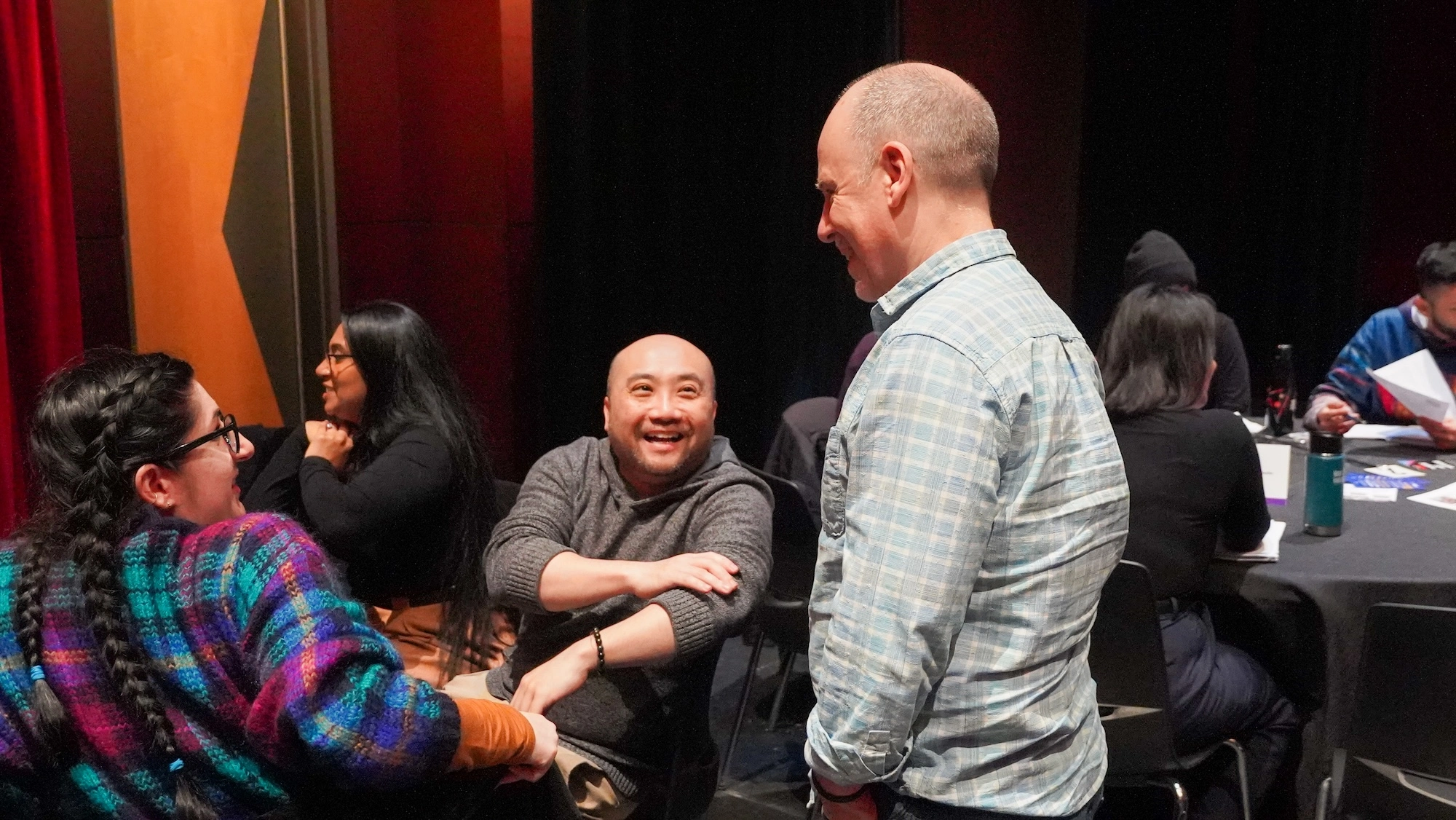 A candid, warm moment between three people at a table in a dimly lit room with red curtains. A man in a grey sweater is laughing heartily while looking up at a man in a blue plaid shirt standing next to him. A candid, warm moment between three people at a table in a dimly lit room with red curtains. A man in a grey sweater is laughing heartily while looking up at a man in a blue plaid shirt standing next to him.