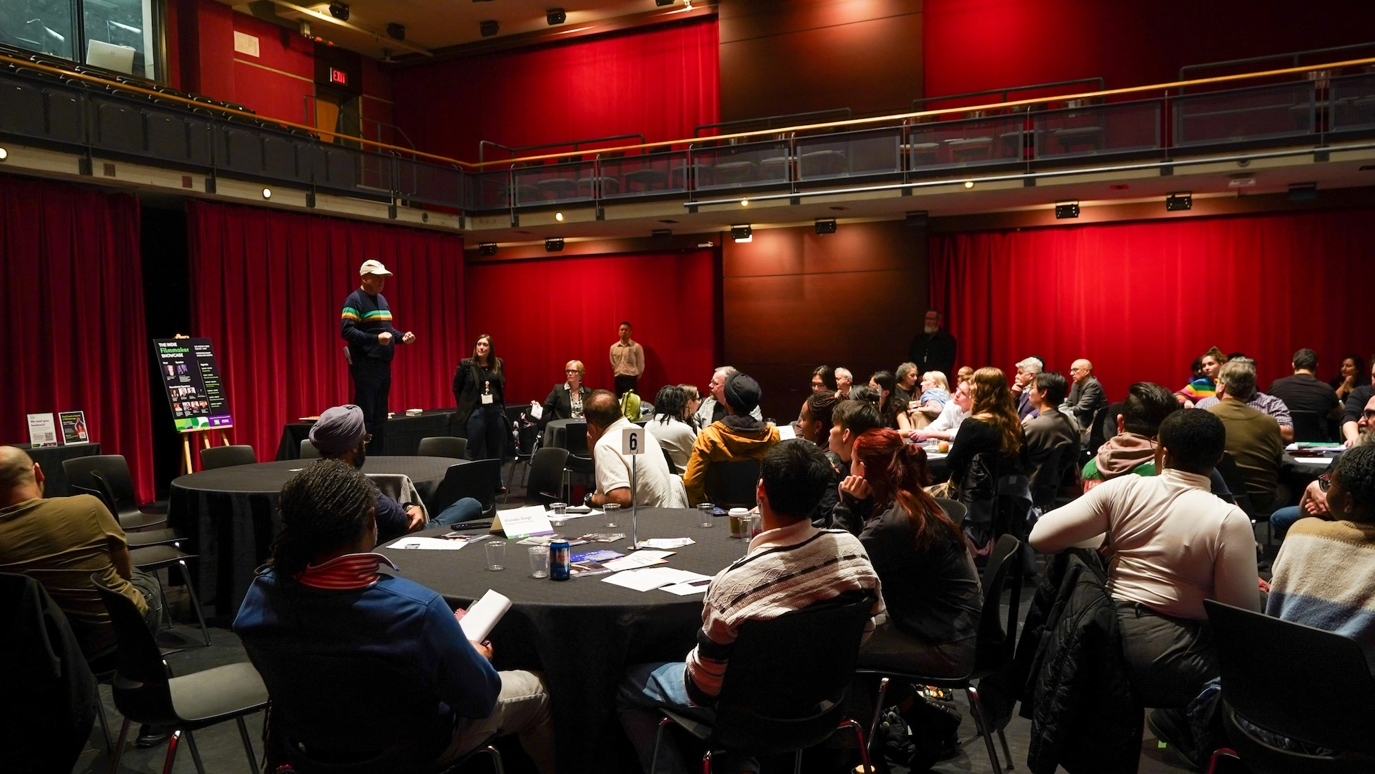 A wide view of a large room with dramatic red floor-to-ceiling curtains. A man in a baseball cap and striped sweater addresses a group of people seated at round tables covered in black cloths. A sign for "The Indie Filmmakers" is visible on an easel. A wide view of a large room with dramatic red floor-to-ceiling curtains. A man in a baseball cap and striped sweater addresses a group of people seated at round tables covered in black cloths. A sign for "The Indie Filmmakers" is visible on an easel.