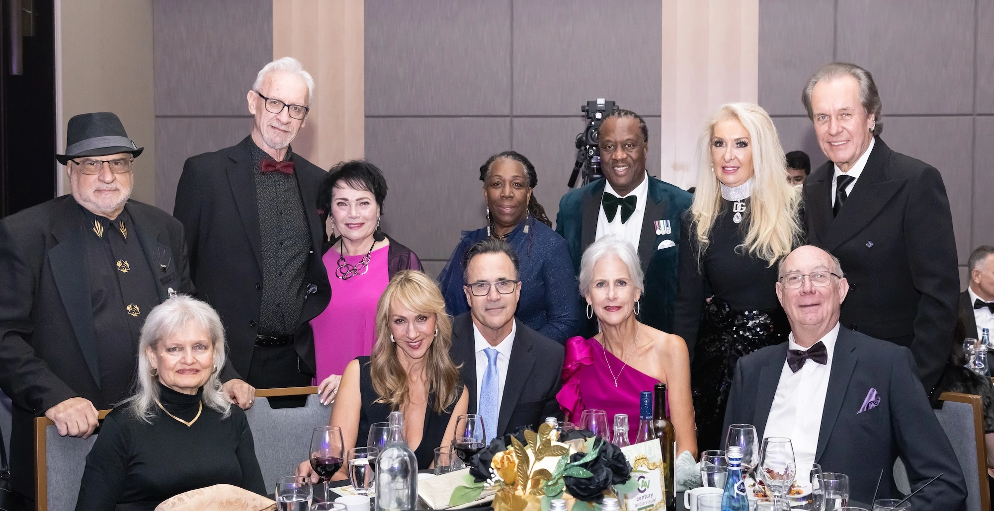 A large group of elegantly dressed guests posing for a photo at a formal gala dinner table, featuring floral centrepieces and wine glasses.