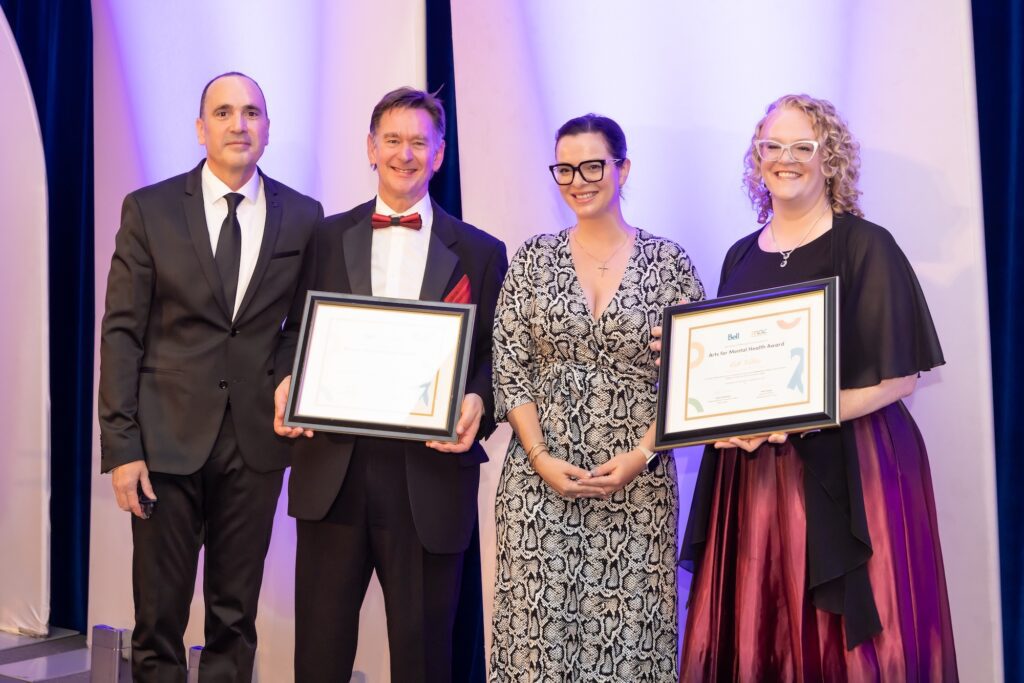 Four people posing on stage at the 2025 MACsquerade Glam Ball. The two individuals in the center and on the right are holding framed "Arts for Mental Health Award" certificates.