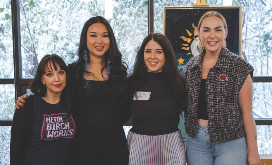 Four women smile together in a brightly lit gallery space with large windows looking out onto snow-covered branches. Behind them is a piece of artwork with a night sky and star motifs.