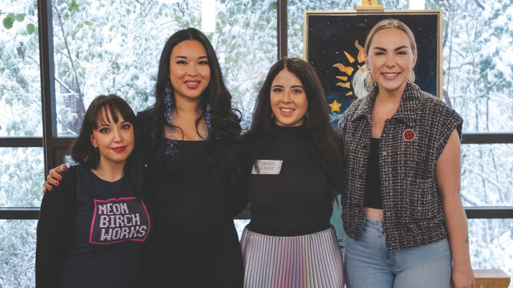 Four women smile together in a brightly lit gallery space with large windows looking out onto snow-covered branches. Behind them is a piece of artwork with a night sky and star motifs.