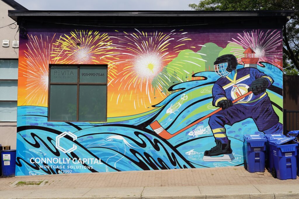 A wide shot of a vibrant, colourful mural on the side of a building. The mural features a hockey player in a blue and yellow "Port Credit Hockey Association" jersey skating on stylized blue waves. The background is filled with fireworks a small lighthouse sits on the horizon. In the bottom left corner, white text reads "Connolly Capital Mortgage Solutions."